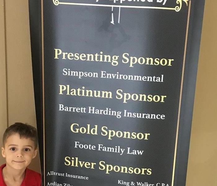 Young boy in red shirt standing in front of black banner listing sponsors